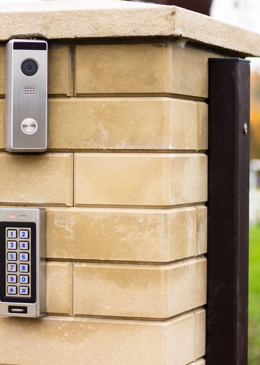 Silver intercom call panel with blue number buttons and a video camera on a brick beige fence post of a private house